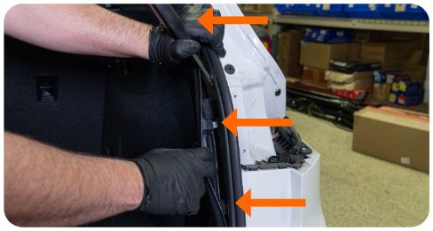 Hands using a wedge tool to pull the trunk liner back, with three arrows marking the metal tabs