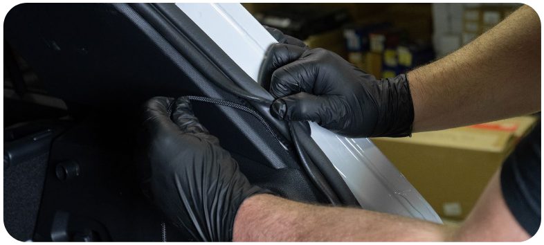 Gloved hands lifting the weather strip to route the tail light harness through the hatch wiring loom on a MK8 Golf