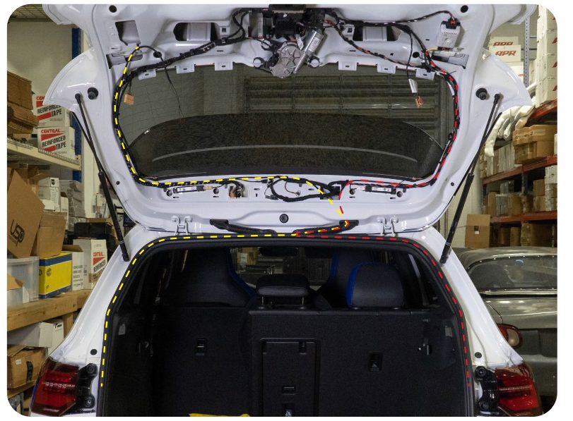 Rear of a MK8 Golf with dashed yellow and red lines tracing the harness route from the quarter panel tail lights up through the hatch to the inner tail lights